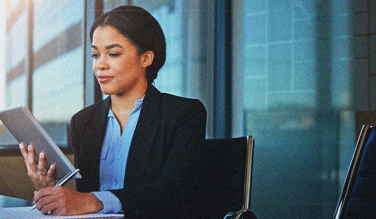 A woman in business attire sits at a desk, holding a tablet in one hand and a pen in the other, looking at the screen thoughtfully in a modern office setting with large windows.